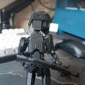K&ouml;nnte beinhalten: Eine schwarze, 3D-gedruckte, bewegliche Actionfigur eines Soldaten, komplett mit einer Ushanka-M&uuml;tze mit Stern, K&ouml;rperpanzerung und einem Sturmgewehr. Die Figur ist so positioniert, als w&auml;re sie bereit f&uuml;r den Einsatz. Die Figur steht auf einem Schreibtisch.