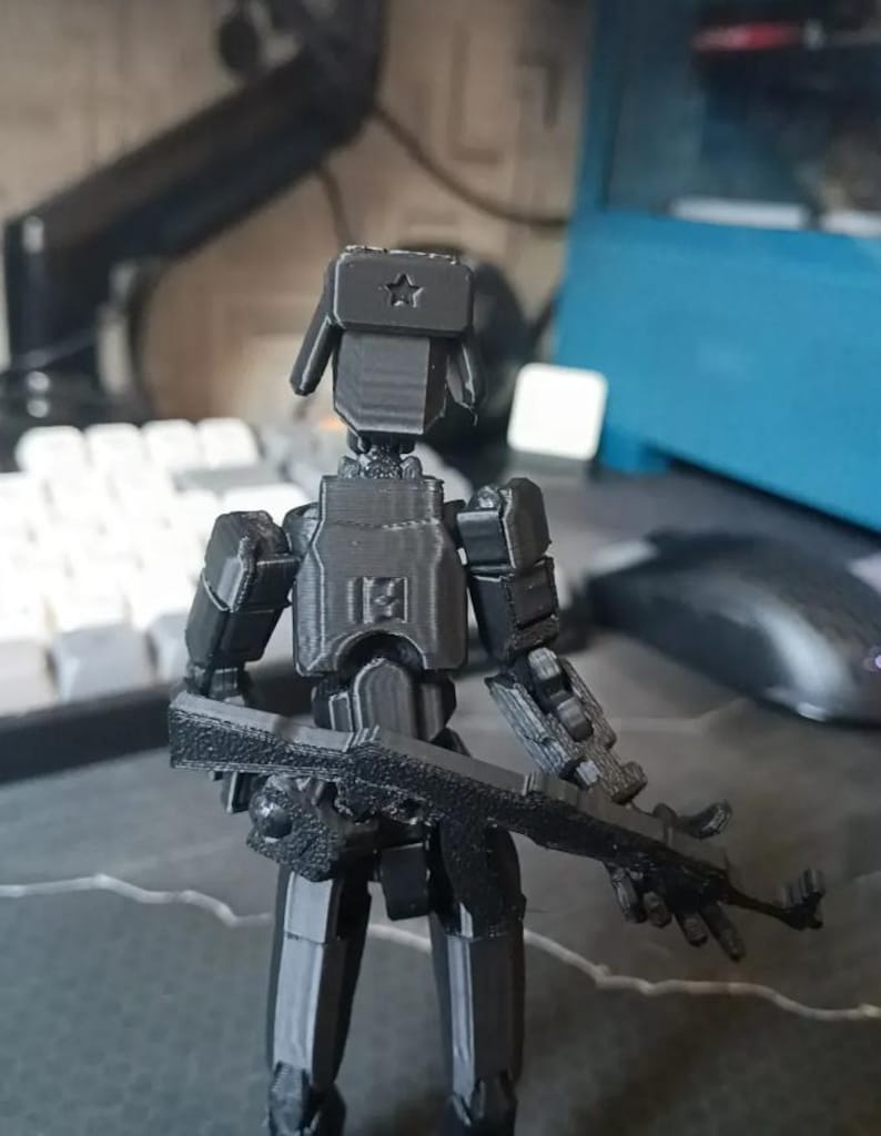 K&ouml;nnte beinhalten: Eine schwarze, 3D-gedruckte, bewegliche Actionfigur eines Soldaten, komplett mit einer Ushanka-M&uuml;tze mit Stern, K&ouml;rperpanzerung und einem Sturmgewehr. Die Figur ist so positioniert, als w&auml;re sie bereit f&uuml;r den Einsatz. Die Figur steht auf einem Schreibtisch.