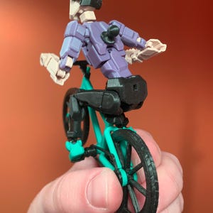 May include: A purple and black articulated action figure riding a teal and black bicycle. The figure is wearing a black helmet and has a black tail.