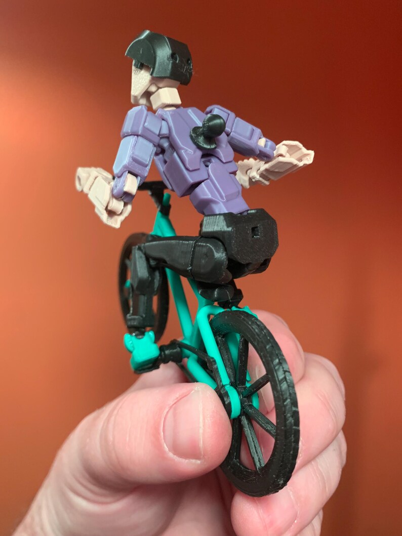 May include: A purple and black articulated action figure riding a teal and black bicycle. The figure is wearing a black helmet and has a black tail.
