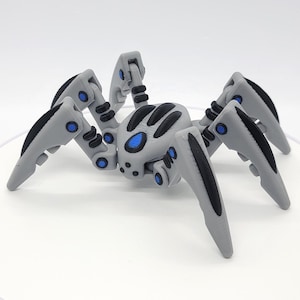 Könnte beinhalten: Ein graues und schwarzes, bewegliches Sp Spielzeug mit blauen Akzenten. Die Spinne hat sechs Beine, einen segmentierten Körper und einen detaillierten Kopf. Die Beine und der Körper sind grau, mit schwarzen Details an Beinen und Körper. Blaue Kreise befinden sich an den Gelenken.