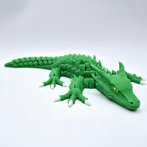 May include: A green articulated dragon figurine with white accents on its claws. The dragon is lying down, with its head and tail visible. The eyes are yellow. The figurine is made of plastic.