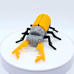 Könnte beinhalten: Ein gelb-graues Spielzeug-Käfer mit schwarzen Beinen und großen gelben Zangen. Der Käfer ist aus Kunststoff und sitzt auf einer weißen Oberfläche. Das Spielzeug ist eine detaillierte Nachbildung eines Käfers.