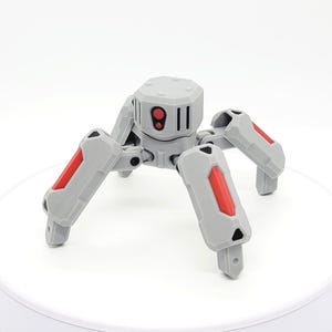 May include: A gray and red toy robot spider with articulated legs and a cylindrical body. The robot has a red eye and vertical black lines on its face. The legs have red accents.