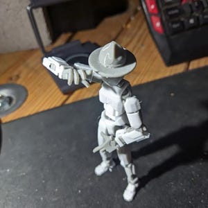 K&ouml;nnte beinhalten: Eine graue Actionfigur eines Soldaten mit Hut und Gewehr. Die Figur steht auf einer dunklen Oberfl&auml;che, im Hintergrund befinden sich eine Tastatur und andere Objekte. Die Figur befindet sich in einer dynamischen Pose.