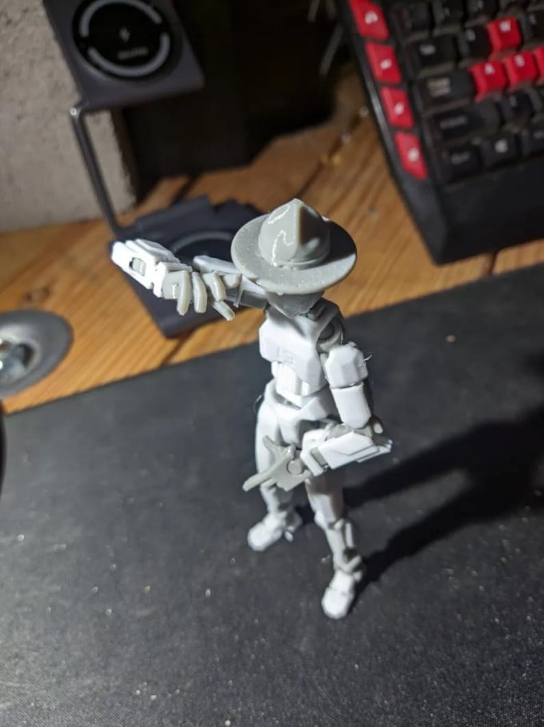 K&ouml;nnte beinhalten: Eine graue Actionfigur eines Soldaten mit Hut und Gewehr. Die Figur steht auf einer dunklen Oberfl&auml;che, im Hintergrund befinden sich eine Tastatur und andere Objekte. Die Figur befindet sich in einer dynamischen Pose.
