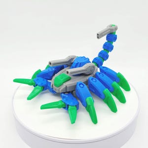 Pode incluir: Um brinquedo de escorpião azul, verde e cinza. O brinquedo tem um corpo segmentado, pernas articuladas e uma cauda curva. O corpo do escorpião é principalmente azul, com detalhes verdes nas pernas e na cabeça. O brinquedo é feito de plástico.