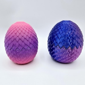 May include: Two decorative, egg-shaped objects with a textured, scale-like pattern. One is a gradient of pink to purple, the other blue to purple. The eggs are separated into two halves, with a jagged edge where they open.