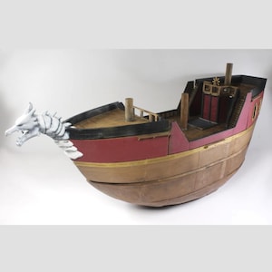 Op de afbeelding: Modelschip met een drakenfiguur. Het schip heeft een rode en gouden romp, houten dekken en een gedetailleerd interieur. De draak is wit met ingewikkelde details. Het schip heeft een uniek, door fantasie geïnspireerd ontwerp.