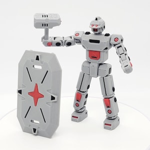 Op de afbeelding: Een grijs en rood gearticuleerd robotspeelgoed met een schild en een wapen. De robot heeft een blokkerig ontwerp met rode accenten op de gewrichten en borst. Het schild is grijs met een rode ster en diagonale lijnen.
