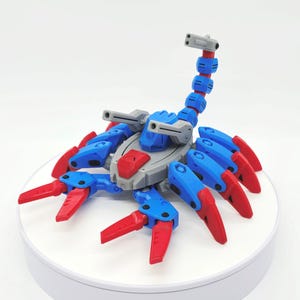 Pode incluir: Um brinquedo de escorpião articulado azul, vermelho e cinza. O brinquedo apresenta um corpo segmentado, garras e uma cauda curva. O corpo do escorpião é principalmente azul com detalhes em vermelho e cinza. O brinquedo é projetado para ser posável.