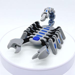 Pode incluir: Uma figura de escorpião cinza e preto com detalhes azuis. O escorpião tem pernas articuladas e uma cauda curva. O corpo apresenta detalhes retangulares azuis. A figura é exibida em uma superfície branca.
