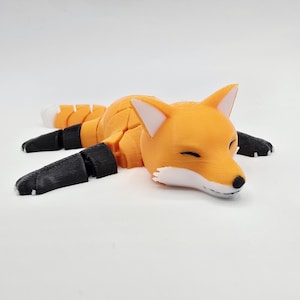 May include: A 3D-printed orange fox figurine with black paws and a white muzzle. The fox is lying down with its eyes closed, suggesting a relaxed pose. The tail is orange and white. The figurine is on a white surface.
