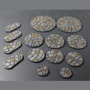 Könnte beinhalten: Eine Sammlung von Miniatur-Pflasterstein-Basen in verschiedenen Formen und Größen. Die Basen sind hauptsächlich grau mit tan- und braunfarbenen Akzenten, die an Kopfsteinpflaster erinnern sollen. Das Set enthält runde und ovale Formen, geeignet für Tabletop-Spiele oder Modellbau.