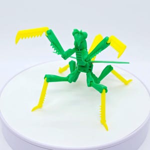 Pode incluir: Um brinquedo de plástico verde e amarelo de louva-a-deus. O inseto tem um corpo segmentado, olhos grandes e patas dianteiras serrilhadas. O brinquedo está posicionado em uma superfície branca, mostrando seu design detalhado e cores vibrantes.
