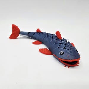 Puede incluir: Un juguete de pez articulado azul y rojo. El pez tiene un cuerpo segmentado con aletas y cola rojas. El pez tiene una cara sonriente con ojos negros y labios rojos. El juguete está diseñado para ser flexible y posable.