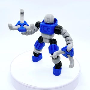 Könnte beinhalten: Ein blaues, graues und schwarzes, bewegliches Roboterspielzeug. Der Roboter hat einen blauen Torso, Beine und Hände, mit grauen Armen und Kopf. Die Gelenke sind schwarz. Das Spielzeug steht auf einer weißen Oberfläche.