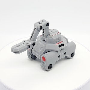May include: A gray toy robot with a movable arm and claw. The robot has red and black accents, with wheels for movement. The design is futuristic and mechanical, suggesting a construction or exploration theme.