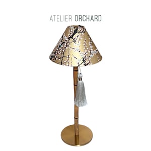 May include: A table lamp with a gold base and a bamboo-style stem. The lampshade has a gold background with a floral pattern in black and white. A silver tassel hangs from the shade. The text "ATELIER ORCHARD" is above the lamp.