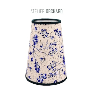 Peut inclure: Abat-jour avec un fond moucheté crème et un motif de feuilles et de branches bleues. Les bords supérieur et inférieur sont bordés d'une bordure vert foncé. Le texte "ATELIER ORCHARD" est imprimé au-dessus de l'abat-jour.