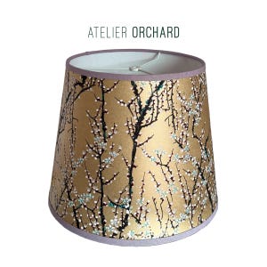 May include: A lampshade with a gold background and a floral design featuring black branches and white and teal blossoms. The lampshade has a lavender trim and the text "ATELIER ORCHARD" at the top.