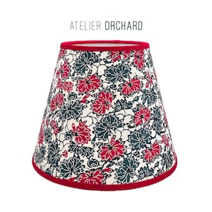 May include: A lampshade featuring a floral design in red and teal on a cream backdrop. The lampshade has a red trim at the top and bottom. The text "ATELIER ORCHARD" is displayed at the top.