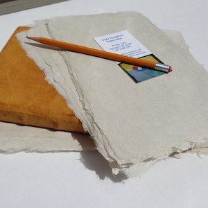 Puede incluir: Una pila de papel hecho a mano con bordes irregulares, un lápiz de madera y una tarjeta de visita. El papel es blanquecino, y el lápiz es naranja con una virola plateada y una goma de borrar rosa. La tarjeta de visita tiene un diseño abstracto colorido.