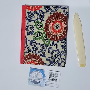 Peut inclure: Un carnet rectangulaire avec une couverture crème ornée d'un motif floral rouge, bleu et vert, doté d'une reliure rouge. À côté se trouve un plioir blanc cassé. Une carte de visite porte le texte Gayle Fitzpatrick Artist/Papermaker.