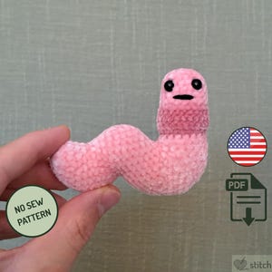 May include: A pink crocheted worm toy with a smiling face. The worm is held in a hand against a light green background. The image includes a small American flag and the text "NO SEW PATTERN".