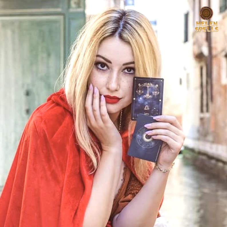 Puede incluir: Una persona con cabello rubio y l&aacute;piz labial rojo sostiene dos cartas del tarot. La carta superior presenta un dise&ntilde;o de balanza de Libra y el s&iacute;mbolo del zodiaco de Libra. La persona lleva una capa con capucha roja. La imagen tiene un fondo borroso con el texto "HELEN SPELLS".