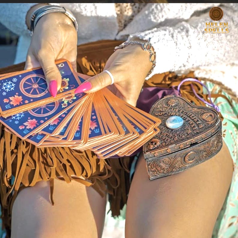 Puede incluir: Un mazo de cartas del tarot con dise&ntilde;os azules y naranjas se abanica, sostenido cerca de una caja en forma de coraz&oacute;n, de color bronce. La caja tiene un dise&ntilde;o decorativo y una piedra azul claro en el centro. El texto "HELEN SPELLS" est&aacute; en la esquina superior derecha.