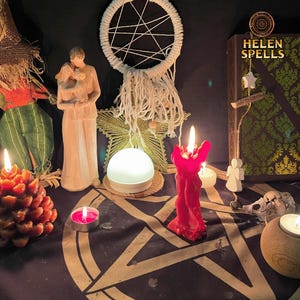 Op de afbeelding: Een rode kaars met een witte droomvanger, twee witte beeldjes en een boek met de titel "Helen Spells" op een zwart oppervlak met een pentagram-ontwerp. Er zijn ook verschillende kleine kaarsen en een dennenappel.