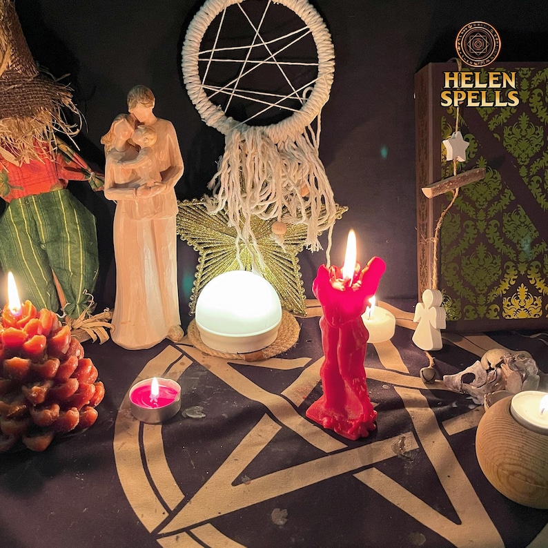 Op de afbeelding: Een rode kaars met een witte droomvanger, twee witte beeldjes en een boek met de titel "Helen Spells" op een zwart oppervlak met een pentagram-ontwerp. Er zijn ook verschillende kleine kaarsen en een dennenappel.