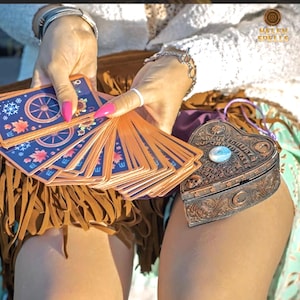 May include: A deck of tarot cards with blue and orange designs is fanned out, held near a heart-shaped, bronze-colored box. The box has a decorative design and a light blue stone in the center. The image includes the text "HELEN SPELLS".