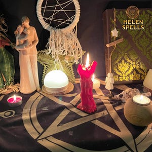 Op de afbeelding: Een rode kaars met een menselijk figuurontwerp is aangestoken op een zwarte altaarbekleding met een pentagramontwerp. Er zijn twee witte kaarsen, een wit licht en een houten doos met de tekst "HELEN SPELLS" erop.