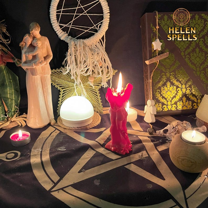 Op de afbeelding: Een rode kaars met een menselijk figuurontwerp is aangestoken op een zwarte altaarbekleding met een pentagramontwerp. Er zijn twee witte kaarsen, een wit licht en een houten doos met de tekst "HELEN SPELLS" erop.