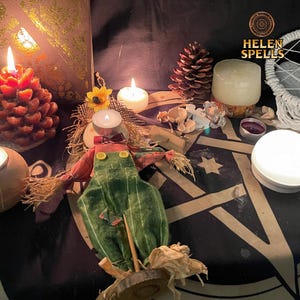 Puede incluir: Un arreglo con una muñeca espantapájaros, velas encendidas y una piña. El espantapájaros lleva un mono verde y una camisa roja. El texto "HELEN SPELLS" es visible. La escena está sobre una superficie oscura con un patrón de estrellas.