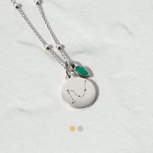 May include: A silver necklace with a round pendant displaying a constellation and a small green gemstone. The necklace has a delicate chain with small ball accents. The pendant has a constellation design.