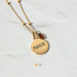 May include: A gold necklace with a circular pendant engraved with the name "PHOEBE". It includes a small, red, teardrop-shaped gemstone and a fine chain with small gold beads. The necklace is a piece of jewellery.