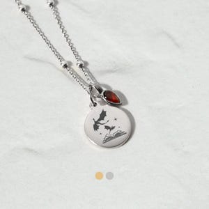 May include: Silver necklace with a round pendant featuring a dragon and book design, and a red gemstone. The chain is delicate with small ball details. A fantasy-themed piece.