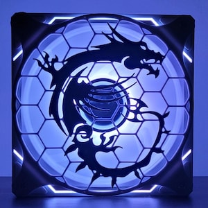 Puede incluir: Un ventilador de ordenador negro con un diseño de panal blanco y una silueta de dragón negro. El ventilador tiene luces LED azules alrededor del borde.