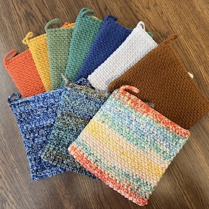 May include: A collection of handmade, knitted pot holders in various colors, including orange, yellow, green, blue, white, and brown. Each pot holder has a loop for hanging. The pot holders are arranged on a wooden surface.
