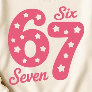 May include: Cream-colored sweatshirt with a pink number 67 design filled with white stars. The word "Six" is above the number 6 and "Seven" is below the number 7. The sweatshirt is a birthday-themed design.