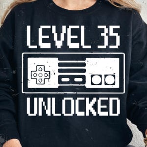 Peut inclure: Sweat-shirt noir avec l'inscription blanche "LEVEL 35 UNLOCKED". Le design comprend une manette de jeu pixélisée, idéal pour un anniversaire ou un passionné de jeux vidéo.