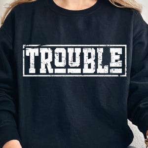 Puede incluir: Sudadera negra con la palabra "TROUBLE" en letras blancas grandes y desgastadas dentro de un marco rectangular blanco. La sudadera es de color liso y tiene cuello redondo.