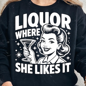May include: Black sweatshirt with a white graphic. The graphic features a retro illustration of a woman holding a martini glass, with the text "LIQUOR WHERE SHE LIKES IT" in a bold, vintage font. The design includes star accents.