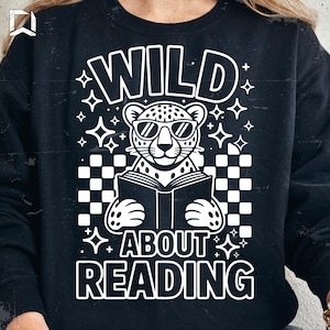 Peut inclure: Sweat-shirt noir avec un motif blanc. Le motif représente un léopard portant des lunettes de soleil et lisant un livre, avec les mots "WILD ABOUT READING" en gras. Le motif comprend des étoiles et un motif à carreaux.