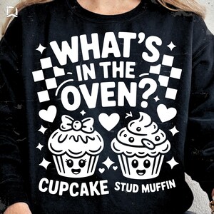 Peut inclure: Sweatshirt noir avec un motif blanc. Le motif comprend le texte "WHAT'S IN THE OVEN?", deux illustrations de cupcakes, des cœurs, des étoiles et l'inscription "CUPCAKE STUD MUFFIN".