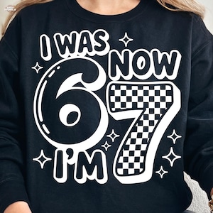 Peut inclure: Sweat-shirt noir avec l'inscription blanche "I WAS 6 NOW I'M 7". Le chiffre 7 présente un motif à carreaux. Le motif comprend des étoiles.