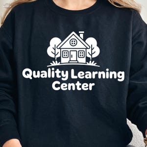 Könnte beinhalten: Schwarzes Sweatshirt mit weißer Grafik eines Hauses, Bäumen und dem Schriftzug "Quality Learning Center". Das Design ist mittig platziert. Das Sweatshirt ist aus weichem Material.